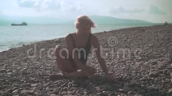 年轻漂亮的性感女孩穿着泳衣坐在岩石海岸上穿着泳衣的金发女郎坐在岸边视频的预览图