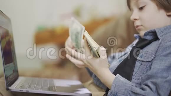 那个穿着牛仔裤夹克的小男孩坐在沙发上手提电脑前数着钱一捆美元的儿童视频的预览图