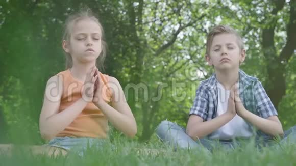 漂亮的小女孩和坐在草地上沉思的英俊男孩孩子们在瑜伽精神发展视频的预览图