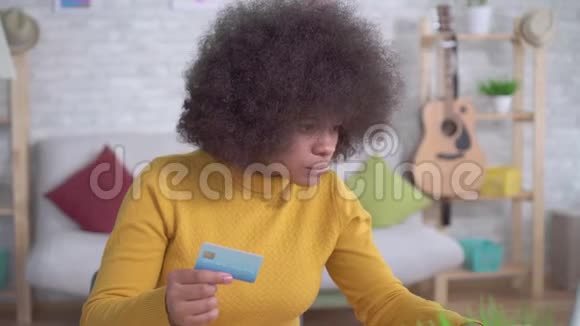 漂亮的非裔美国女孩发型是非洲式的手里拿着银行卡现代公寓里拿着笔记本电脑视频的预览图