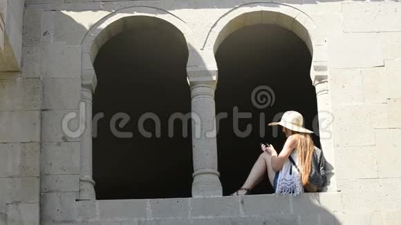 戴着背包的帽子的年轻女人自拍坐在老窗的拱门里夏日阳光明媚旅费视频的预览图
