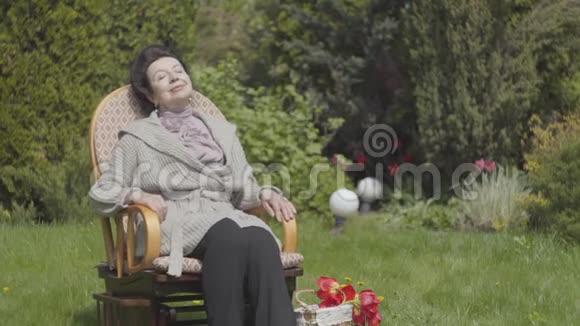 肖像可爱快乐成熟的女人坐在摇椅上享受阳光有郁金香的篮子视频的预览图