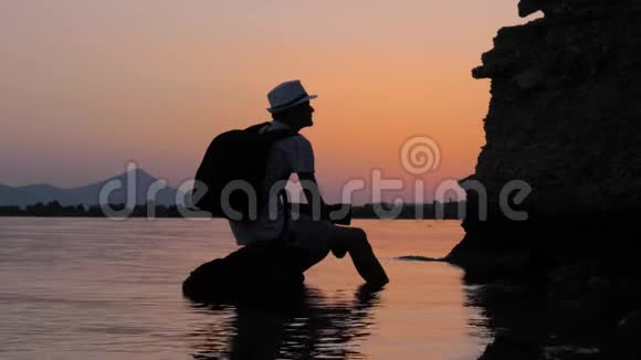 游客在海中的石头上休息黄昏时分希普斯特坐在海中的岩石上戴帽子的男性视频的预览图