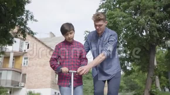 肖像可爱的年轻父亲教他的儿子在公园里骑滑板车他支持孩子帮助他视频的预览图