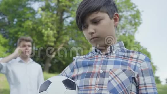 一个悲伤的男孩看着镜头把足球放在前台年轻忙碌的父亲在说话视频的预览图