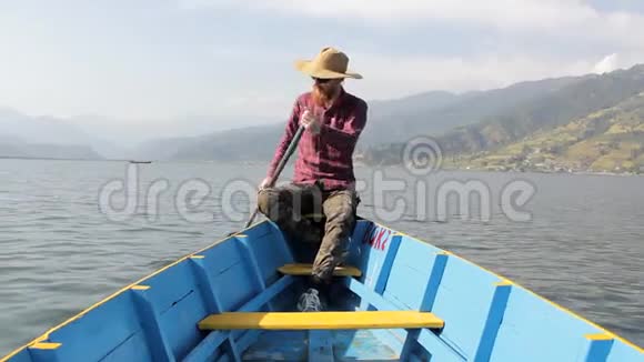 大胡子穿着红色衬衫戴着帽子戴着墨镜穿着蓝色的木船在湖边划桨背景是群山视频的预览图