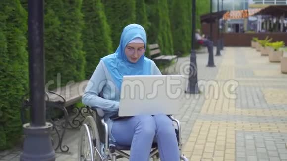 戴着传统围巾的年轻穆斯林妇女使用坐在轮椅上的笔记本电脑视频的预览图