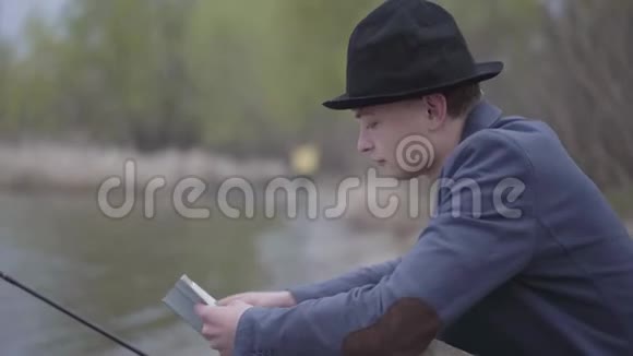 一位穿着夹克衫戴着野帽的时尚渔夫坐在河边看着一本书或钓鱼提示视频的预览图