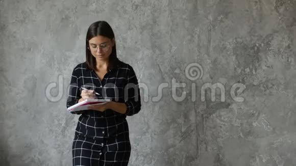 一个年轻快乐的女孩在一个灰色的墙上写笔记本时髦的年轻女子视频的预览图