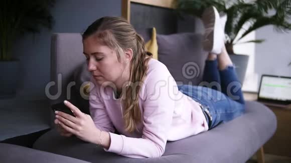 漂亮的女孩穿着粉红色毛衣在客厅里躺在沙发上用智能手机打字看录像视频的预览图