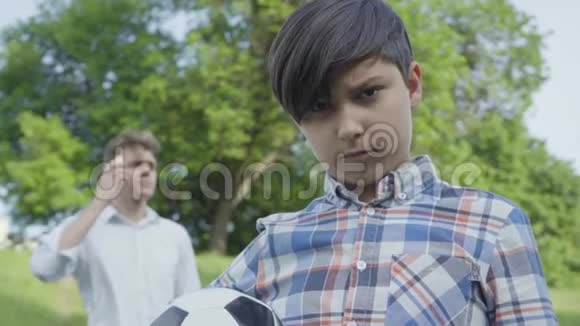 一个悲伤的男孩看着镜头把足球放在前台年轻忙碌的父亲在说话视频的预览图