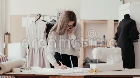 裁缝在工作室里打电话在布料上画一支铅笔在背景中有一个衣架视频的预览图