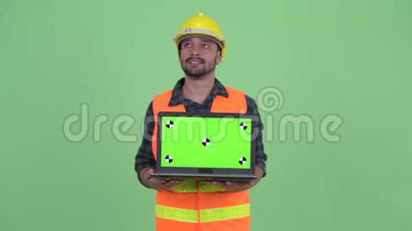 快乐的年轻有胡子的波斯人建筑工人一边思考一边展示笔记本电脑视频的预览图