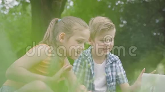 公园里一台笔记本电脑的镜头中一个可爱的英俊男孩和一个长发的漂亮女孩挥舞着手的肖像视频的预览图