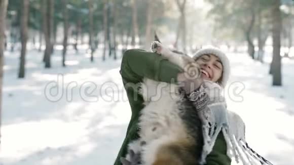 快乐的女人戴着羊毛帽戴着长长的温暖围巾在寒冷的阳光明媚的日子在下雪的冬天公园里抚摸她的宠物快乐视频的预览图