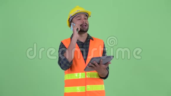 快乐的年轻胡子波斯人建筑工人使用电话和数字平板电脑视频的预览图