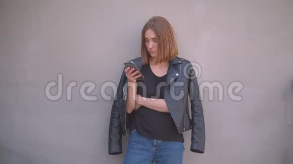 穿着皮夹克的年轻可爱的白种人女孩的特写肖像用手机看室外的摄像机视频的预览图