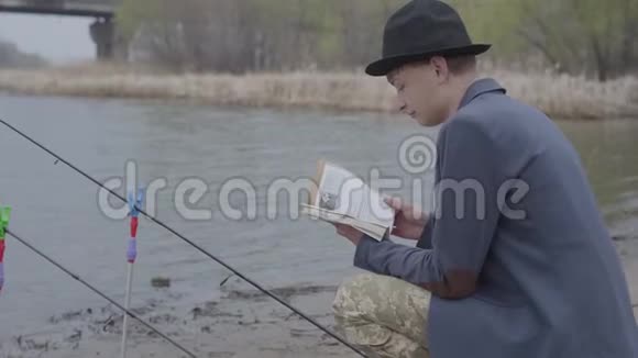 一位穿着夹克衫戴着野帽的时尚渔夫坐在河边看着一本书或钓鱼提示视频的预览图