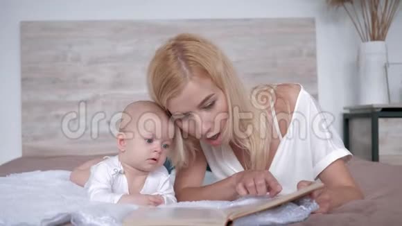微笑的年轻妈妈读着一本书给她的小女儿听她一边好奇地躺在家里的床上视频的预览图