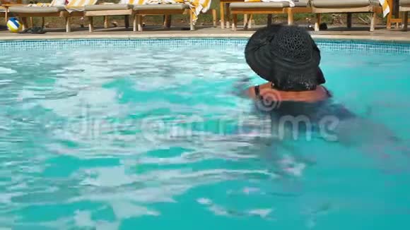 未被识别的高加索老年妇女戴着黑色帽子在酒店的蓝色游泳池游泳在这样的背景下视频的预览图