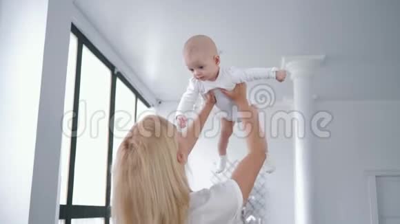 母爱快乐的年轻妈妈把可爱的新生女儿从头上旋转亲吻她在白色的房间里视频的预览图
