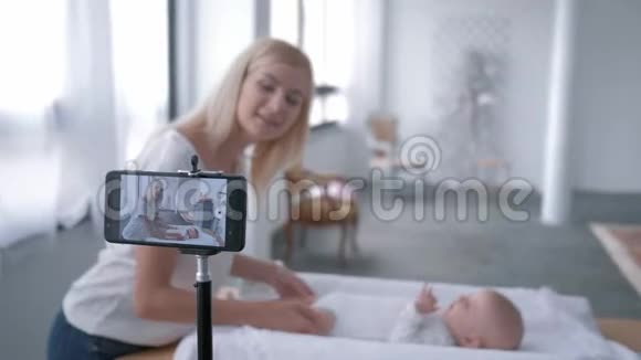 Videoblog年轻的母亲使用手机为躺在桌子上的小女儿做运动视频的预览图