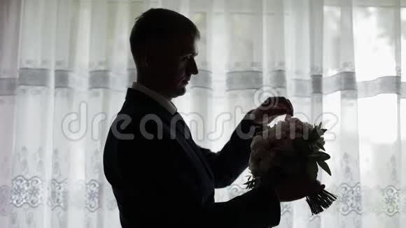 英俊的新郎把他的婚礼花束固定在窗户附近剪影视频的预览图
