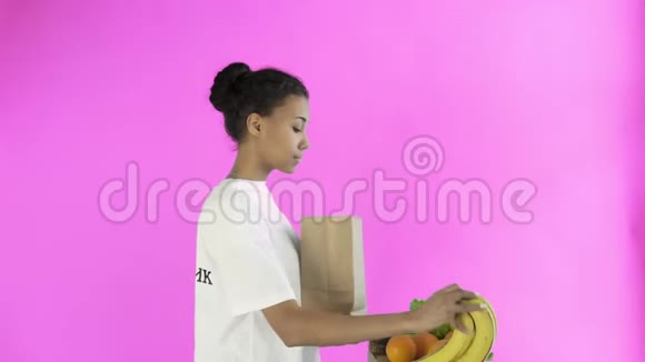 幸福的女人用粉色背景的有机健康蔬菜和水果装入纸袋视频的预览图