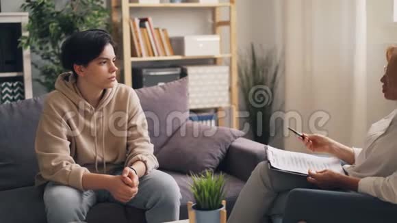 悲伤的十几岁男孩和坐在沙发上的女心理治疗师交谈视频的预览图