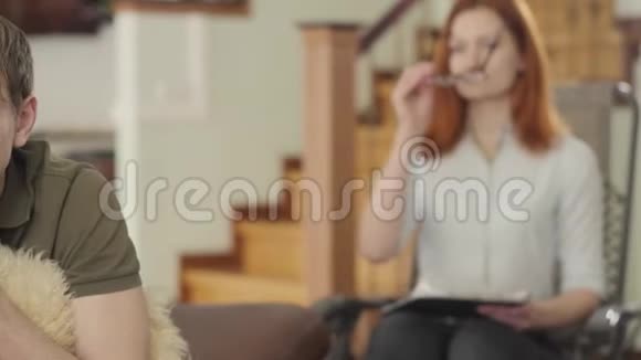 沮丧的年轻人在心理学家的办公室抱着毛茸茸的枕头在前景红发女性视频的预览图