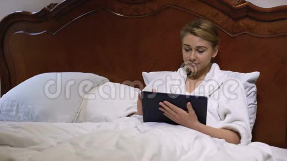 微笑的女人躺在床上在平板电脑上滚动在社交网络上聊天视频的预览图