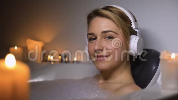 戴着耳机的微笑女孩躺在浴缸里用泡沫和蜡烛看着相机视频的预览图
