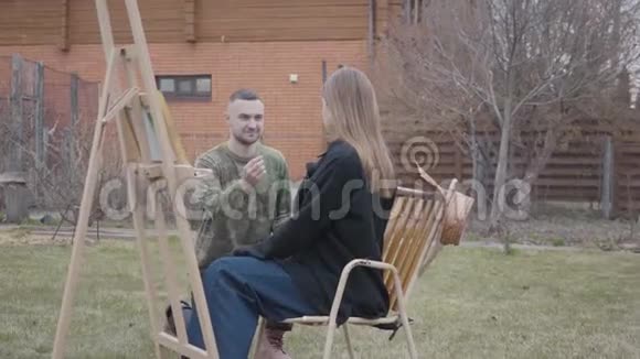 漂亮的年轻女画家坐在户外木制画架前快乐的男人把戒指戴在女士的手指上视频的预览图