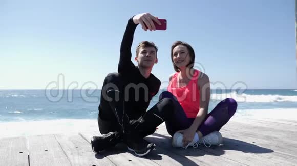 年轻的健康夫妇坐在海滩附近拿着电话摆出自拍姿势快乐的男朋友和女朋友拍照SS视频的预览图