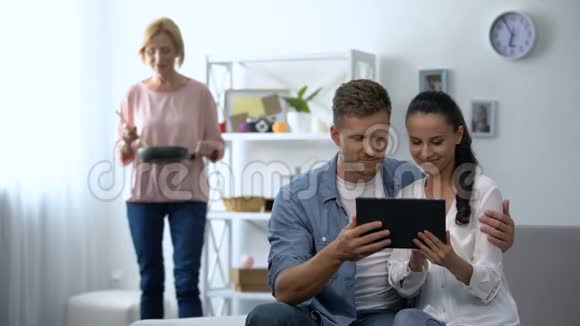 好奇的婆婆看着年轻夫妇在家里的平板电脑上滚动视频的预览图
