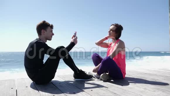 年轻的健康男子坐在海滩上用电话给他的女朋友拍照强烈的波浪在岩石上飞溅视频的预览图