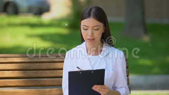 微笑女医生在医院公园的长椅上填写数据在户外工作视频的预览图