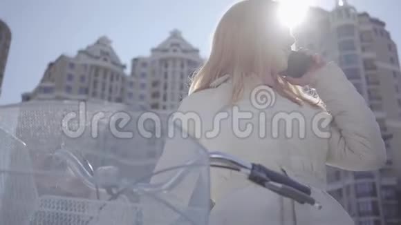 漂亮的金发女人戴着牙套穿着温暖的外套骑着自行车带着篮子用手机说话视频的预览图