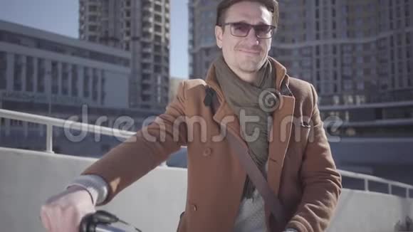 英俊的微笑男人戴着太阳镜和棕色外套骑着自行车站在城市里看着镜头休闲视频的预览图