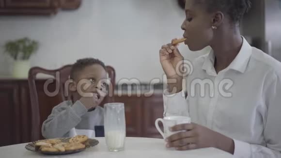 漂亮的非裔美国女人和她的小儿子坐在桌子旁吃饼干和喝牛奶视频的预览图