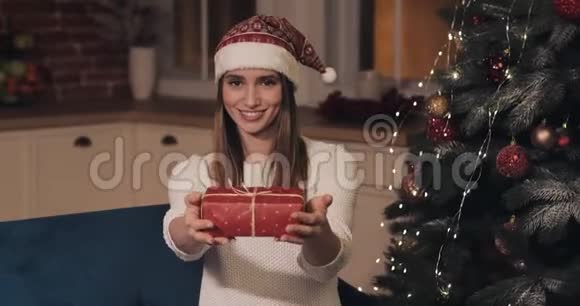 可爱的年轻微笑白人女孩戴着圣诞帽坐在舒适的家圣诞背景送红色礼物视频的预览图