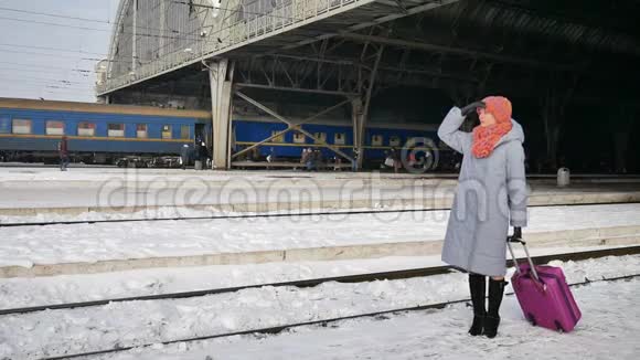 一名身穿蓝色外套头戴彩色帽子的年轻女子在火车站带着紫色行李箱的户外视频视频的预览图