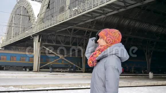 一位穿着蓝色外套和五颜六色帽子的年轻女子在阳光明媚的冬日在火车站的肖像旅程概念视频的预览图