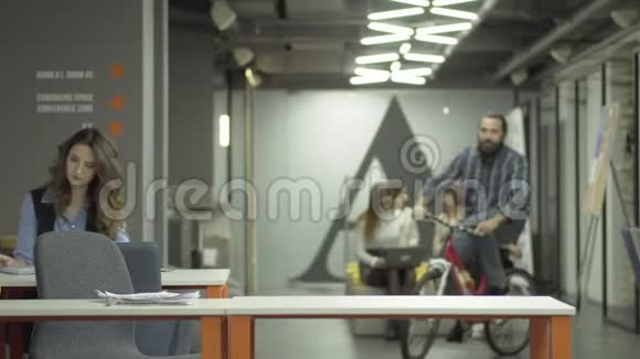 留胡子的微笑男子骑自行车在现代办公室骑士向他的女同事问好坐在他的旁边视频的预览图