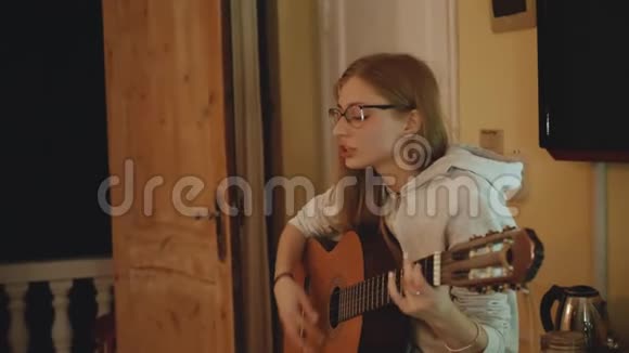 戴眼镜的金发女孩在舒适的公寓里弹吉他在椅子上唱歌一个女人在室内创作音乐视频的预览图
