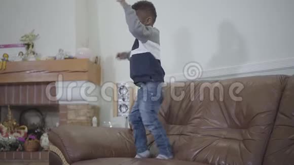 在舒适的客厅背景下非裔美国人可爱的小男孩在沙发上跳跃夫妻关系A视频的预览图