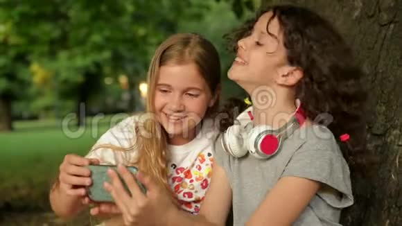 两个可爱的年轻姐妹带着耳机在美丽的日子里玩得开心快乐的孩子们在夏天公园玩看小孩视频的预览图