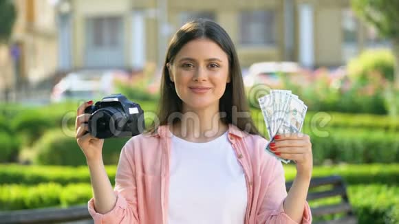 可爱的女性在镜头前展示相机和一堆美元有利可图的职业视频的预览图