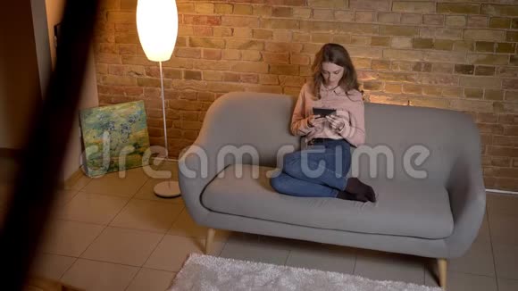 年轻的金发少女坐在沙发上兴致勃勃地看着舒适的家中的平板电脑视频的预览图