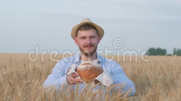 年轻快乐的谷物种植者戴着草帽的帅哥在镜头前给你美味的烘焙面包站在田野里微笑视频的预览图